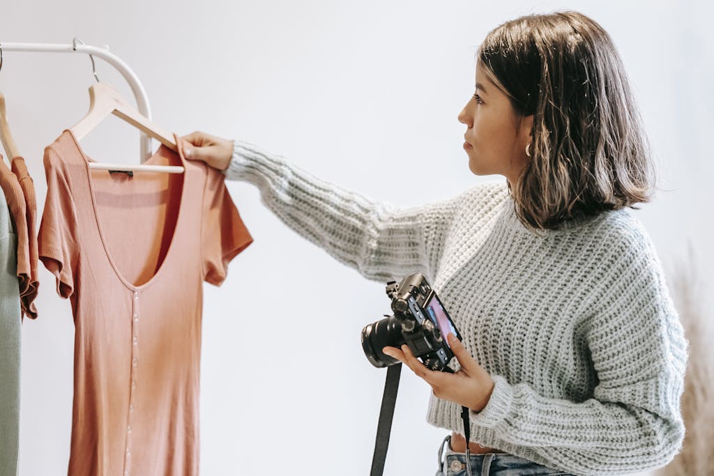 Side view of young female in sweater selecting shirt from hanger for photoshoot while holding camera in light studio